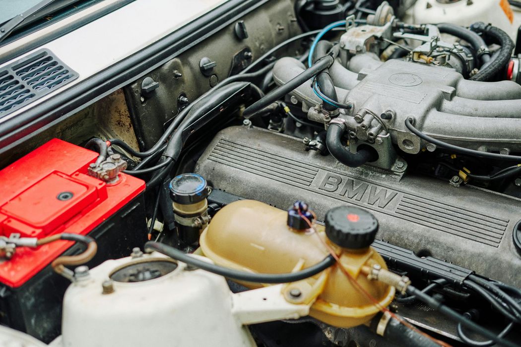 BMW engine bay, with a red battery, beige coolant reservoir, and various hoses and components.