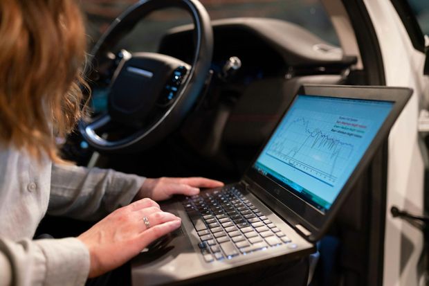 Person using a laptop inside a car, likely for diagnostics. Steering wheel and dashboard visible.