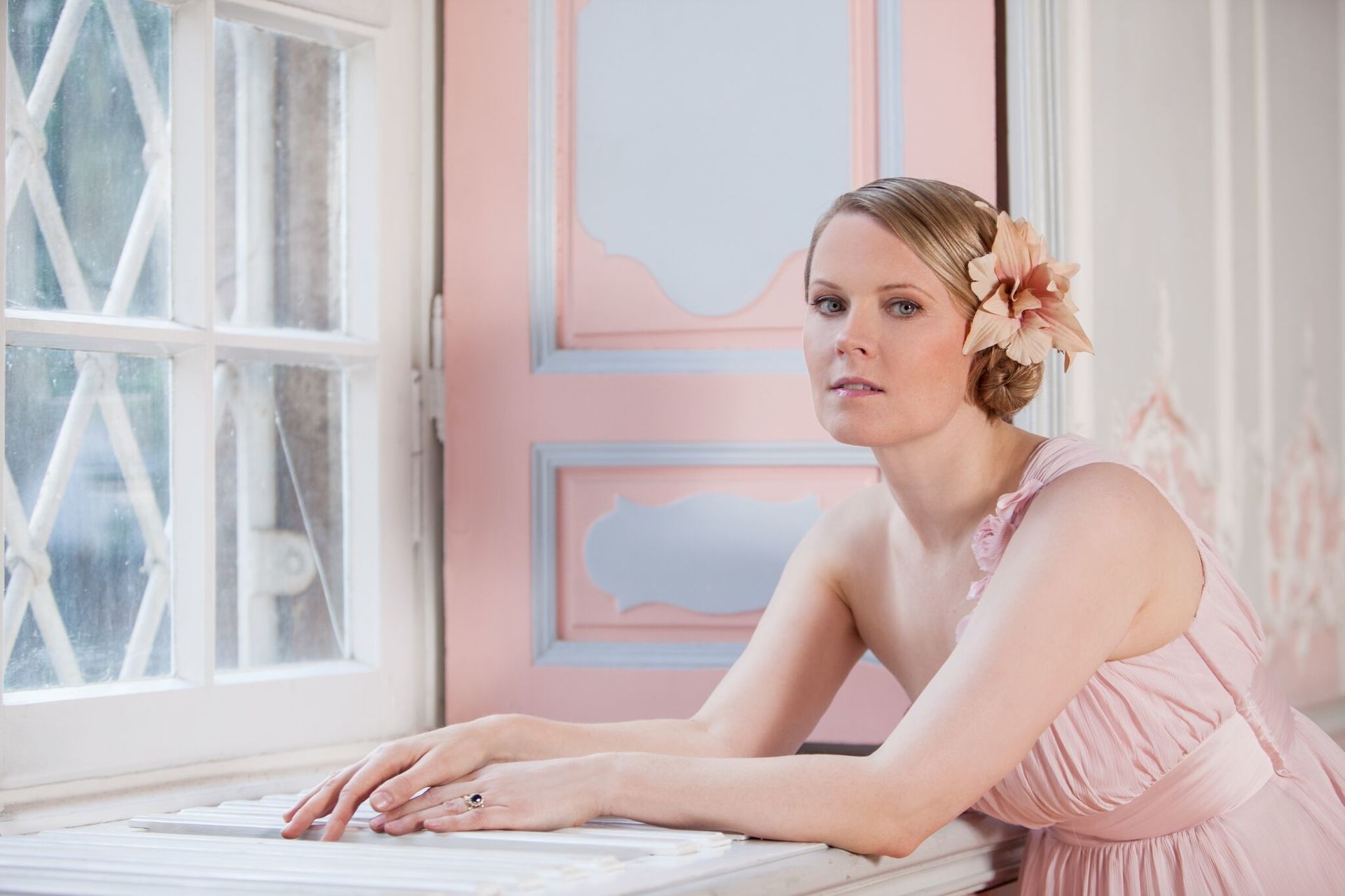 Eine Frau in einem rosa Kleid sitzt mit einer Blume im Haar vor einem Fenster.