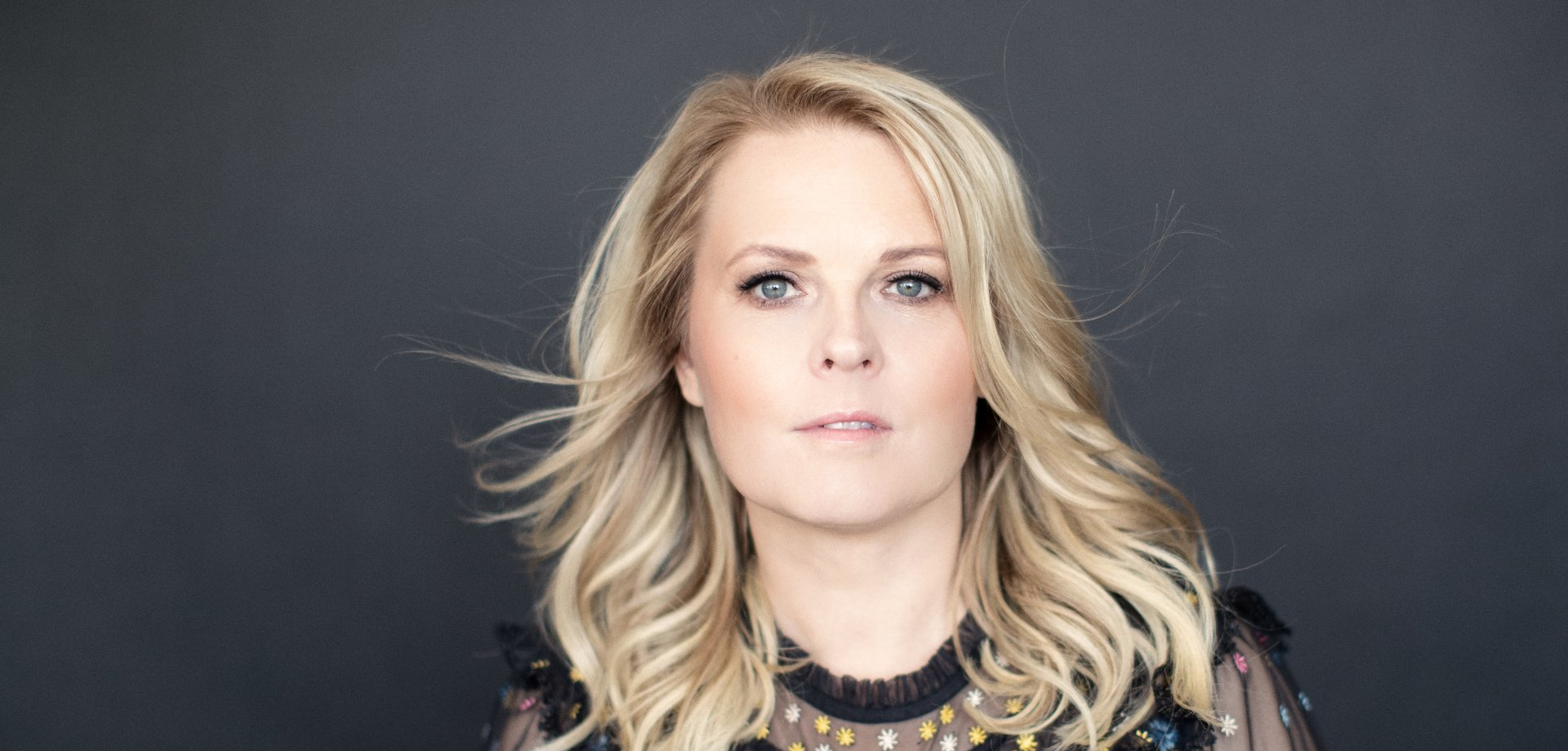 Eine Frau mit blonden Haaren steht vor einem schwarzen Hintergrund und blickt in die Kamera.