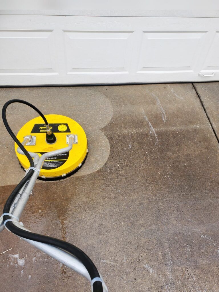 A yellow surface cleaner being used to wash a dirty concrete driveway in front of a white garage door.
