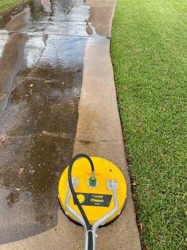 A bright yellow pressure washer surface cleaner sits on a concrete driveway, partially cleaning a dark, stained surface.