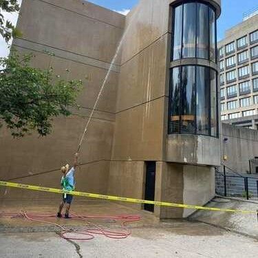 A worker uses a power washer to clean a tall, tan concrete wall next to a glass-paneled building exterior.