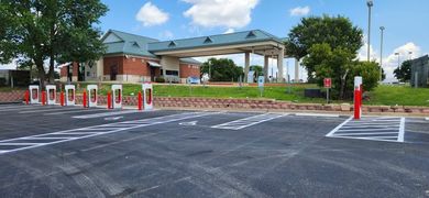 A row of red Tesla Supercharger stations in a paved parking lot with a brick building and green trees in the background.