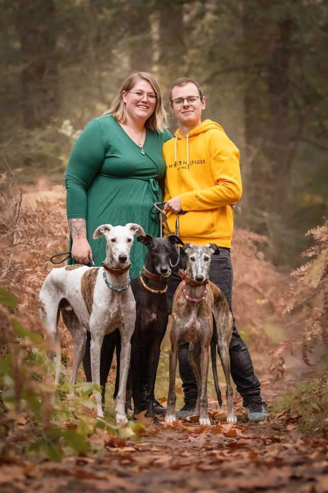 Een persoon in een groene jurk en een ander in een gele hoodie staan ​​in een herfstbos met drie aangelijnde windhonden.