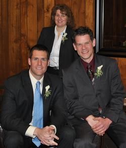 Two men in suits and ties and a woman in a black blazer pose together in front of a wood-paneled wall.