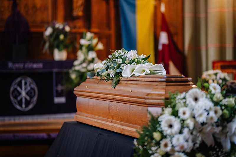 A wooden casket adorned with white flowers sits in a church, with a blue and yellow flag visible in the background.