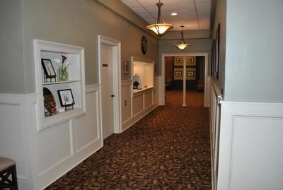 A well-lit hallway with patterned brown carpet, grey walls with white wainscoting, recessed wall displays, and two lamps.