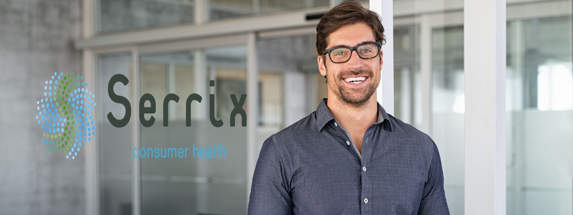 A man wearing glasses is standing in front of a glass door and smiling.