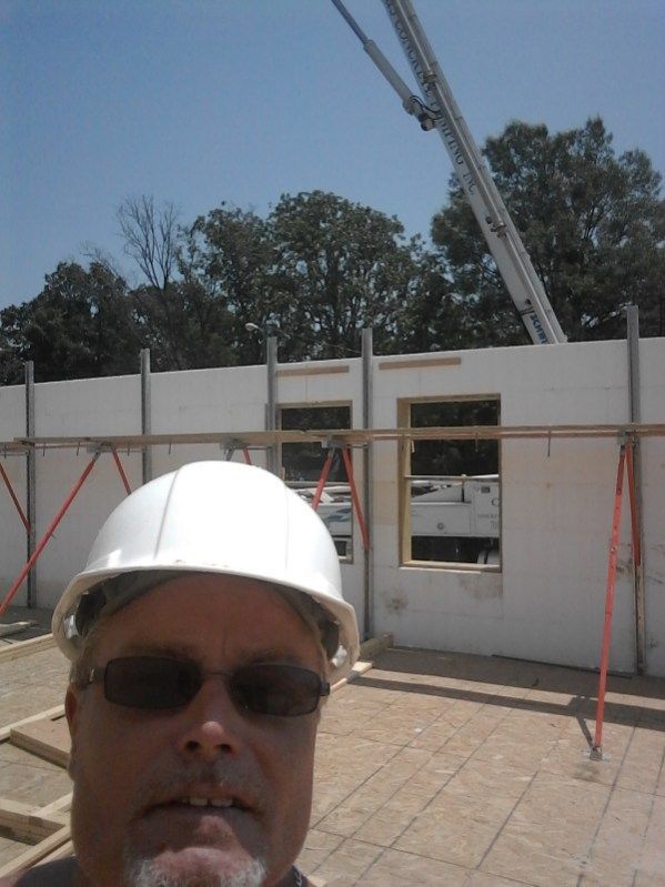 A man wearing a hard hat and sunglasses stands in front of a building under construction