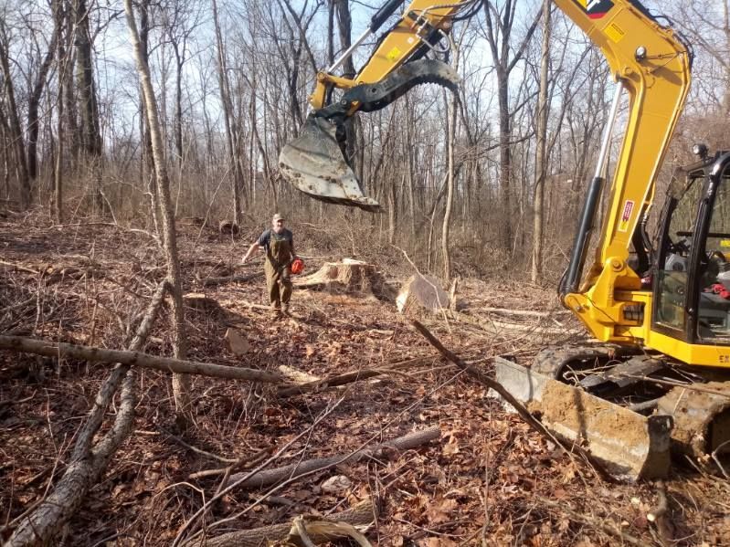 A man is standing next to a yellow excavator in the woods.