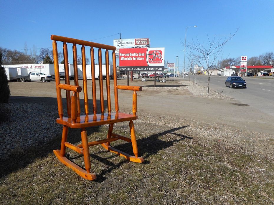 Giant wooden rocking chair on grassy area beside a road, with billboards and a gas station in the background.