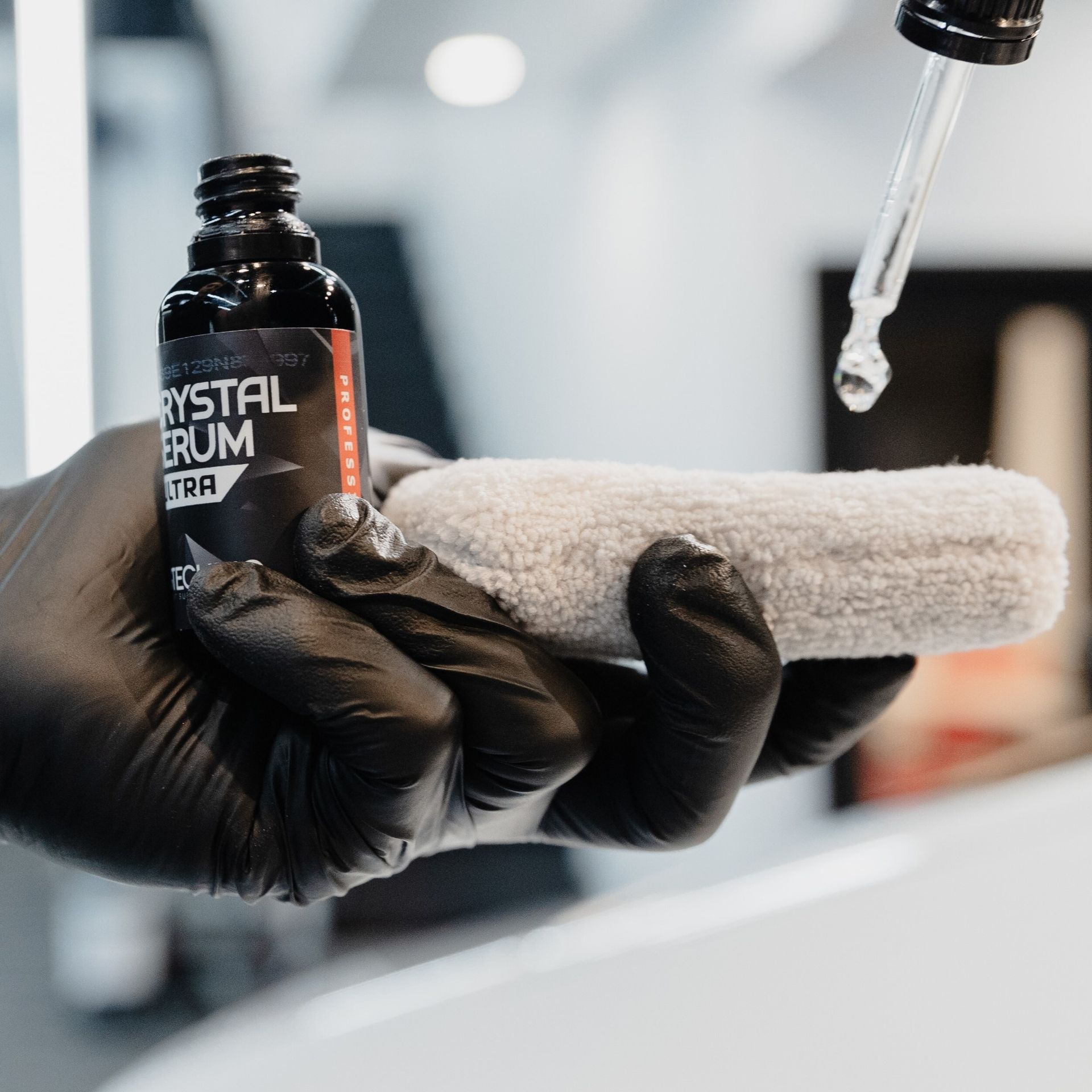 Black-gloved hands hold a serum bottle and white towel under a dropper in a spa-like setting