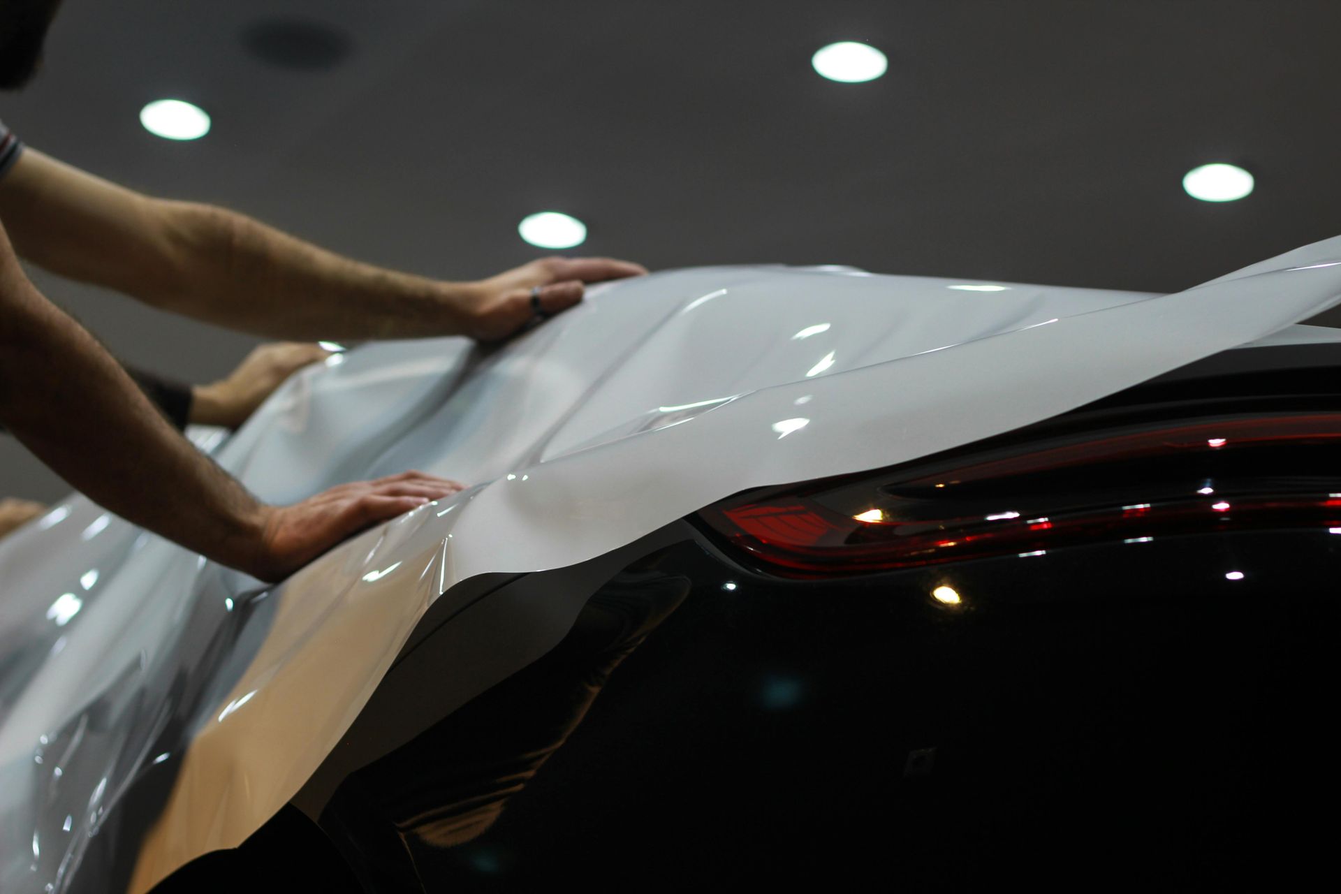 Hands applying white vinyl wrap to a black car under bright showroom lights