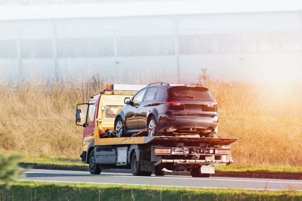 Tow truck hauling a black SUV on a flatbed along a roadside in bright sunlight