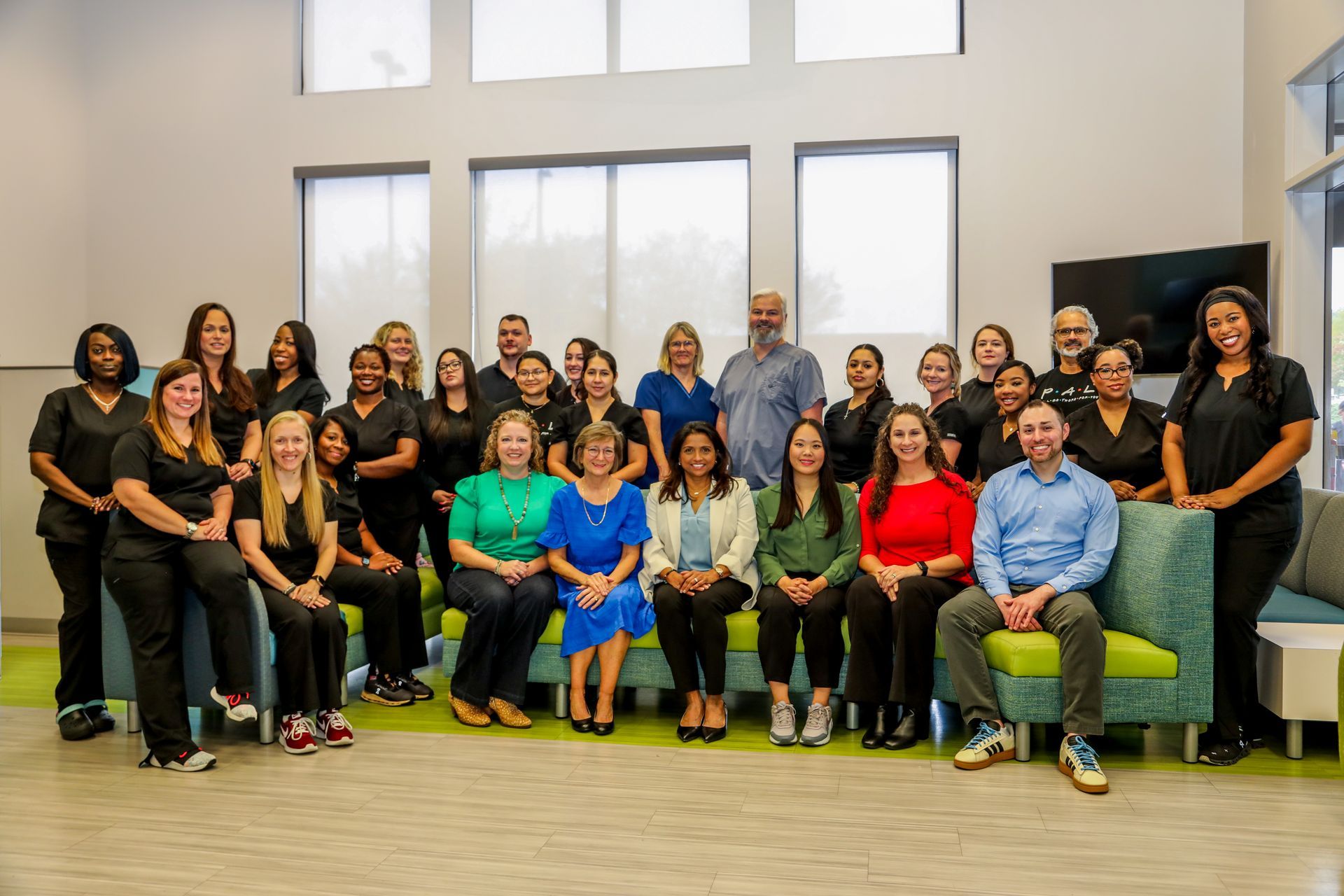Group of people, staff, in a modern office, posing for a group photo; some seated on green and blue seating, smiling.