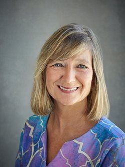 Blond-haired woman smiling, wearing a blue and purple patterned blouse. Gray background.