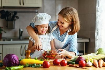 Mother and child cook together in a kitchen, mixing food with vegetables on the counter.