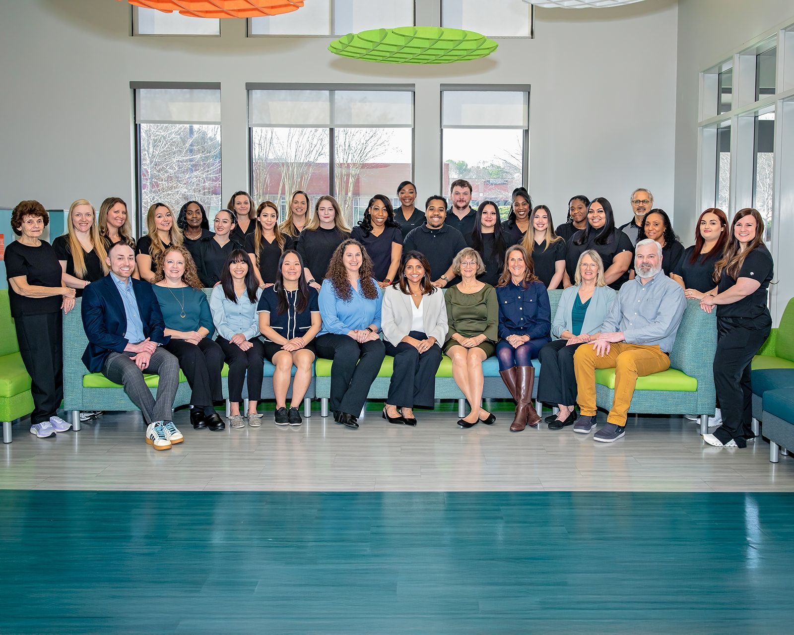Group of people, staff, in a modern office, posing for a group photo; some seated on green and blue seating, smiling.
