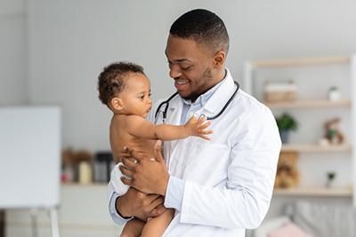Smiling doctor holding a baby in an exam room. The doctor has a stethoscope. The baby wears a diaper.
