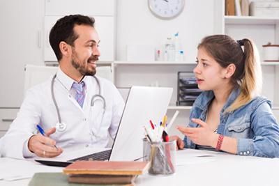 Doctor and patient, a young person, in a medical office, seated, talking, doctor is using a laptop.