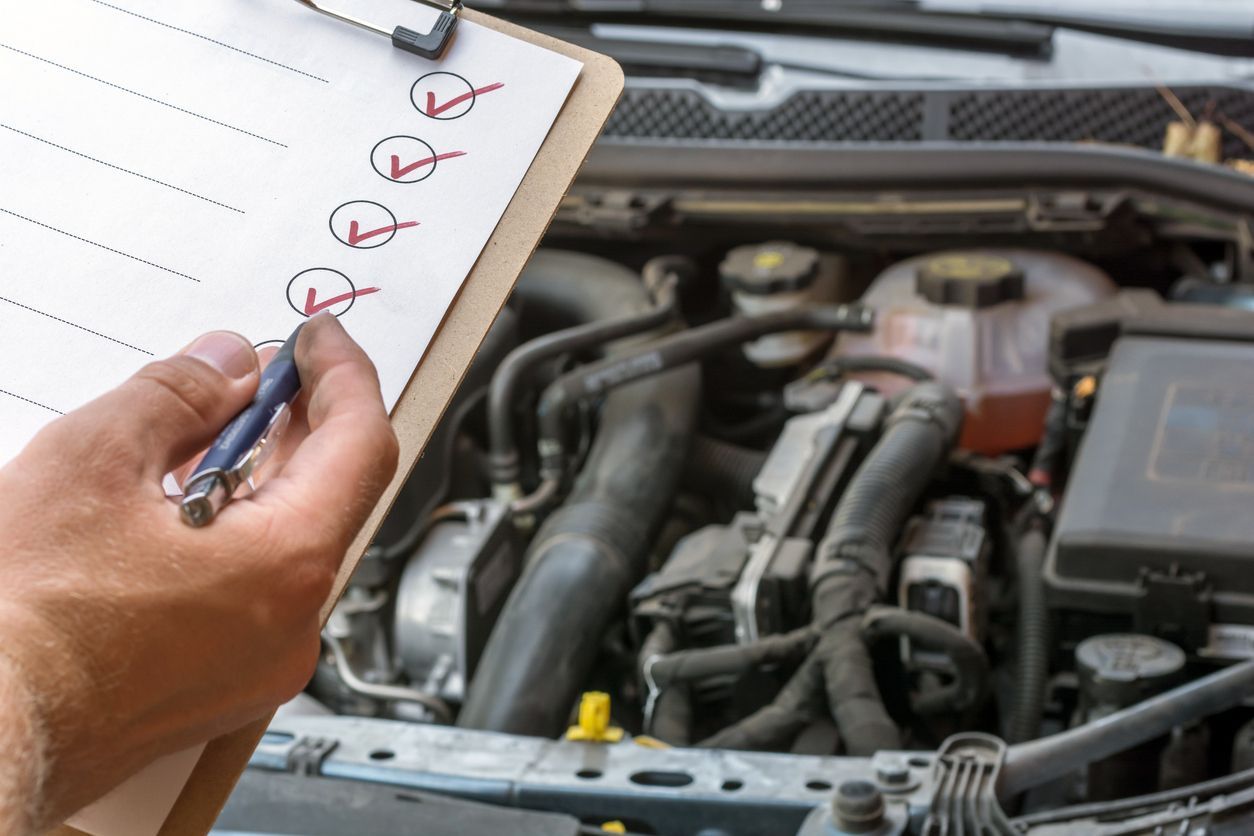 A person is writing on a clipboard in front of a car engine.