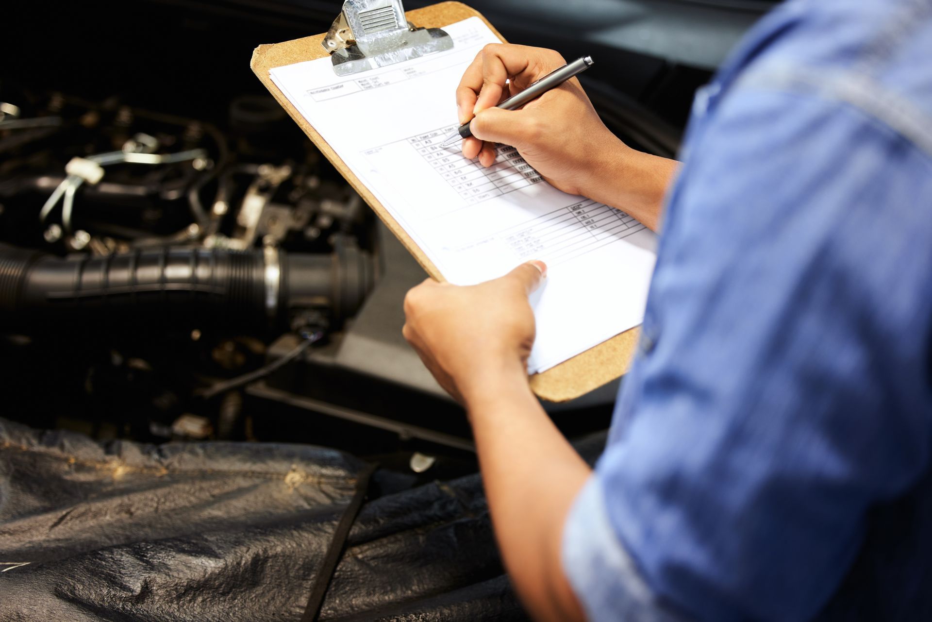 A man is writing on a clipboard in front of a car engine.