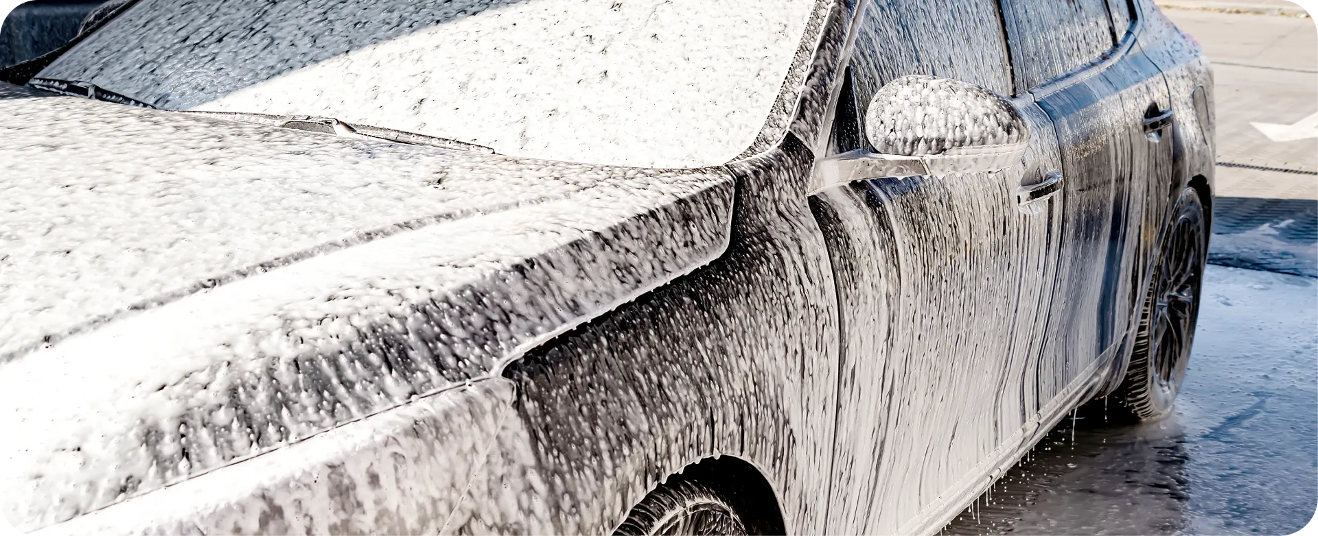A silver sports car covered in white foam, being washed in a driveway.