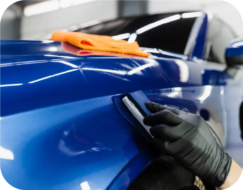 A gloved hand applies ceramic coating to a blue car with a microfiber cloth on the hood.