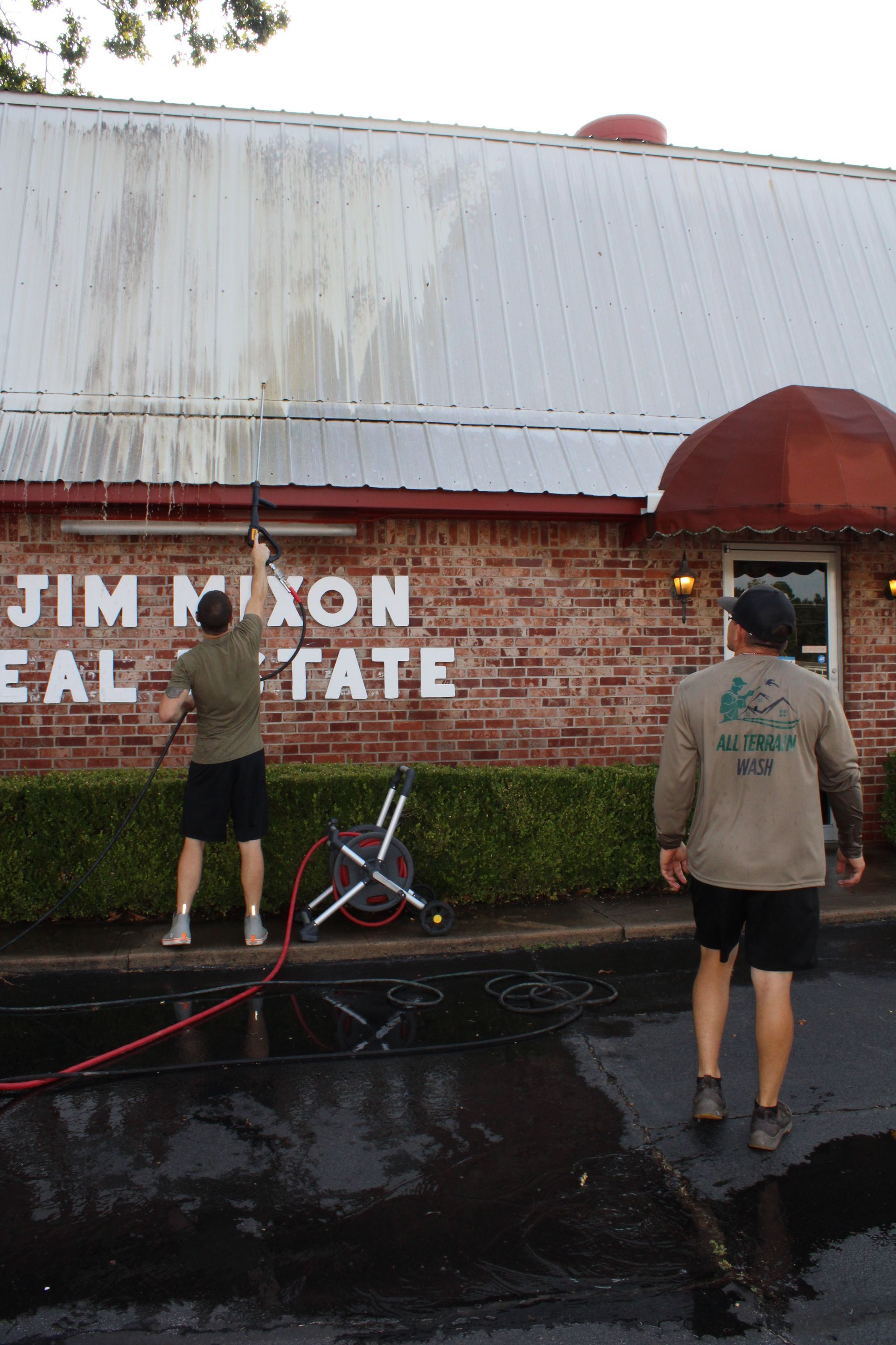 Two men pressure washing a building's metal roof. One holds the wand. Building is brick, with red awnings.