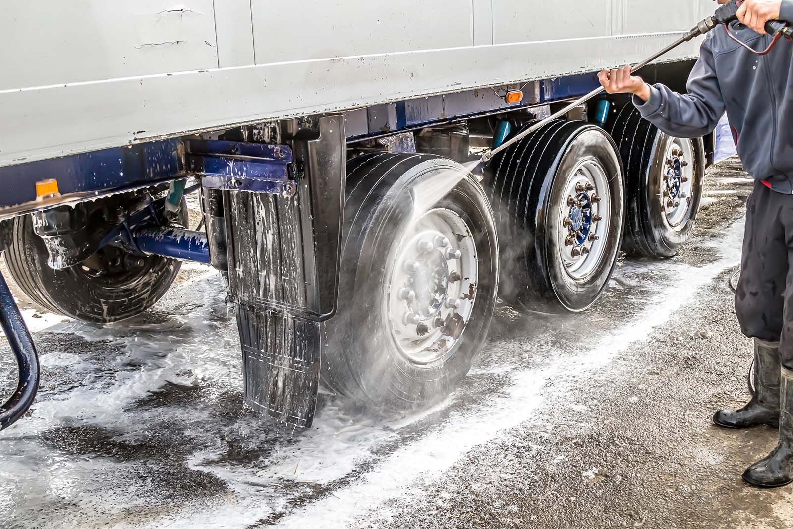Person washing a semi-truck trailer's tires with a pressure washer, spraying water and soap on the wheels.