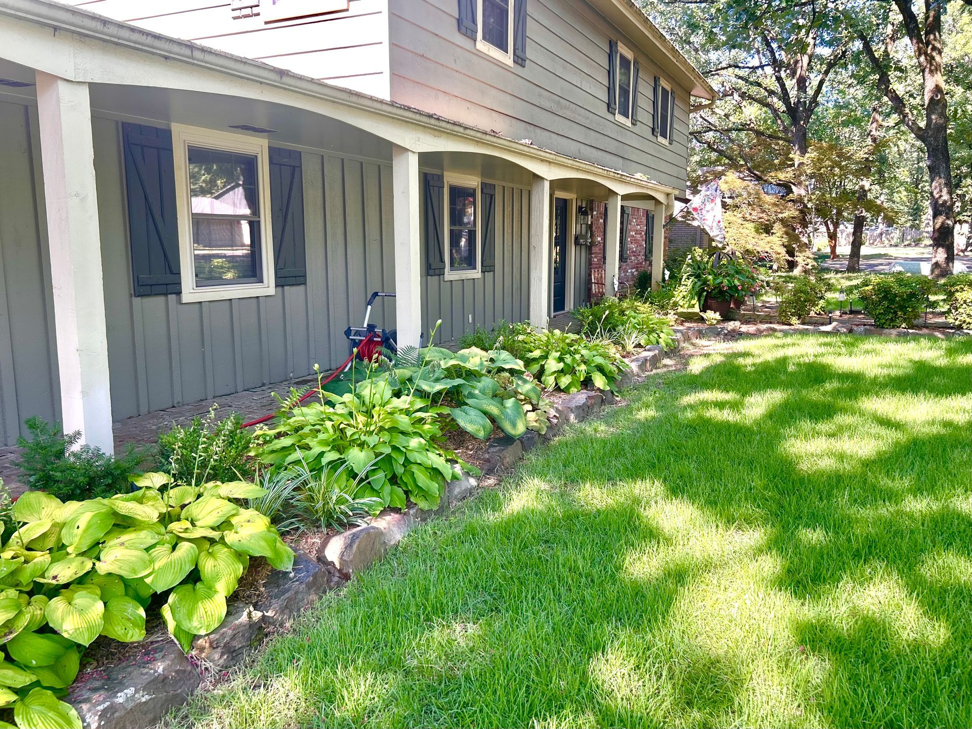 Two-story house with green siding, porch, and green lawn. Hostas line the border of the home.
