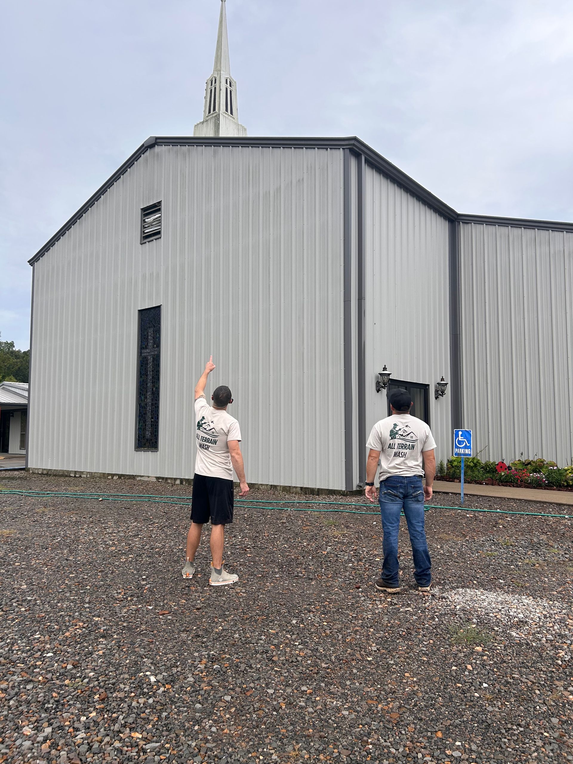 Two men in matching shirts outside a church with one pointing up. Gray building, cloudy sky.