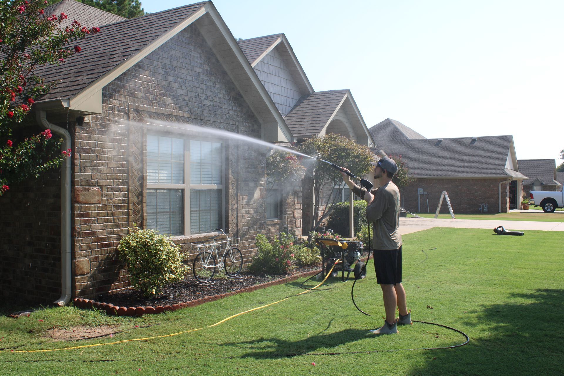 Person power washing the brick facade of a house with a green lawn on a sunny day.
