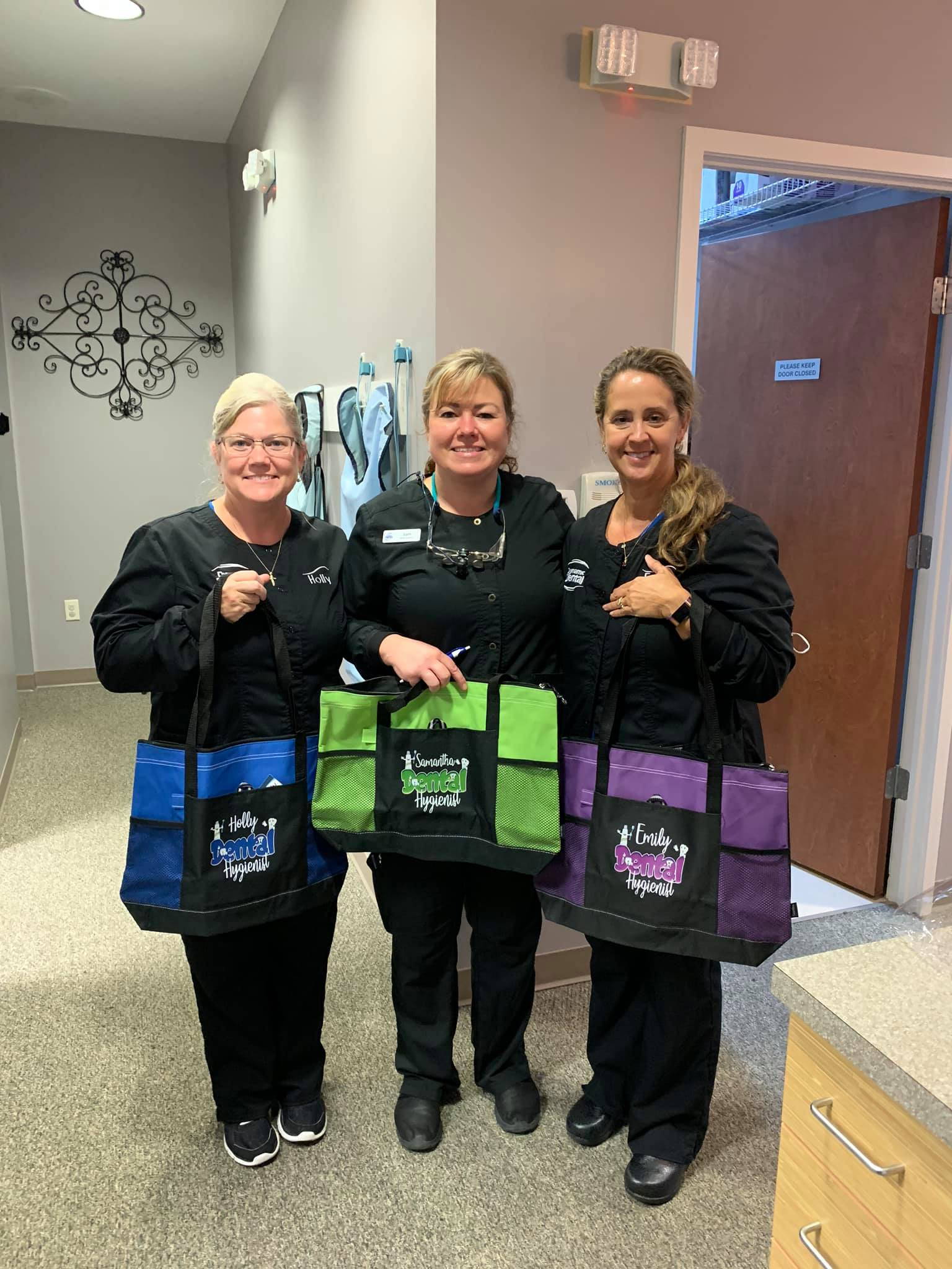 Three dental assistants are standing next to each other in a hallway holding bags.