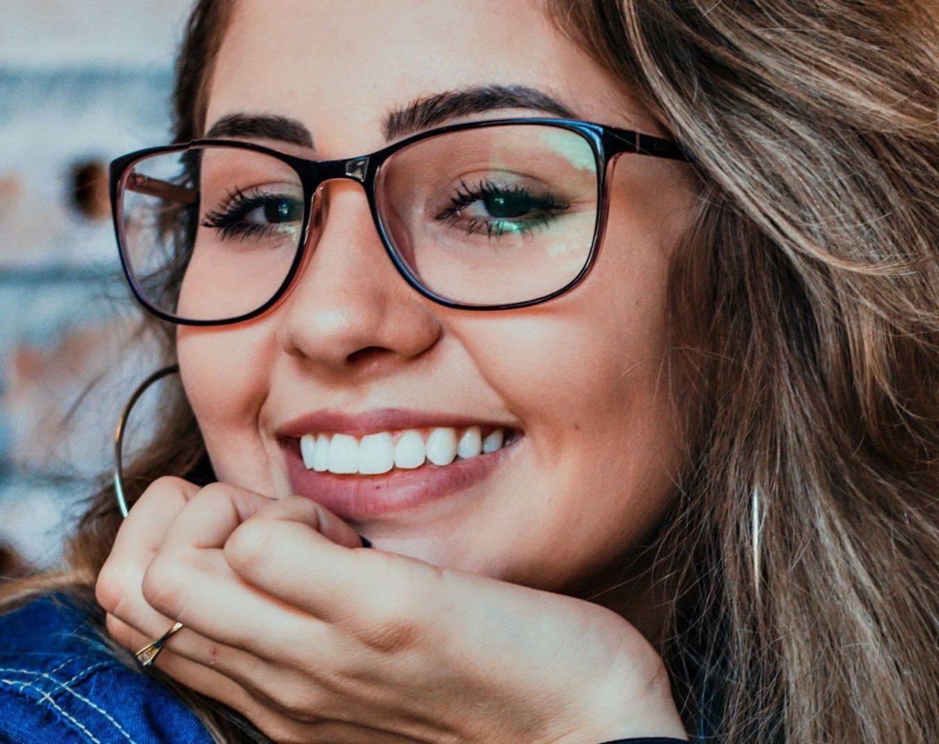 A woman wearing glasses is smiling with her hand on her chin.