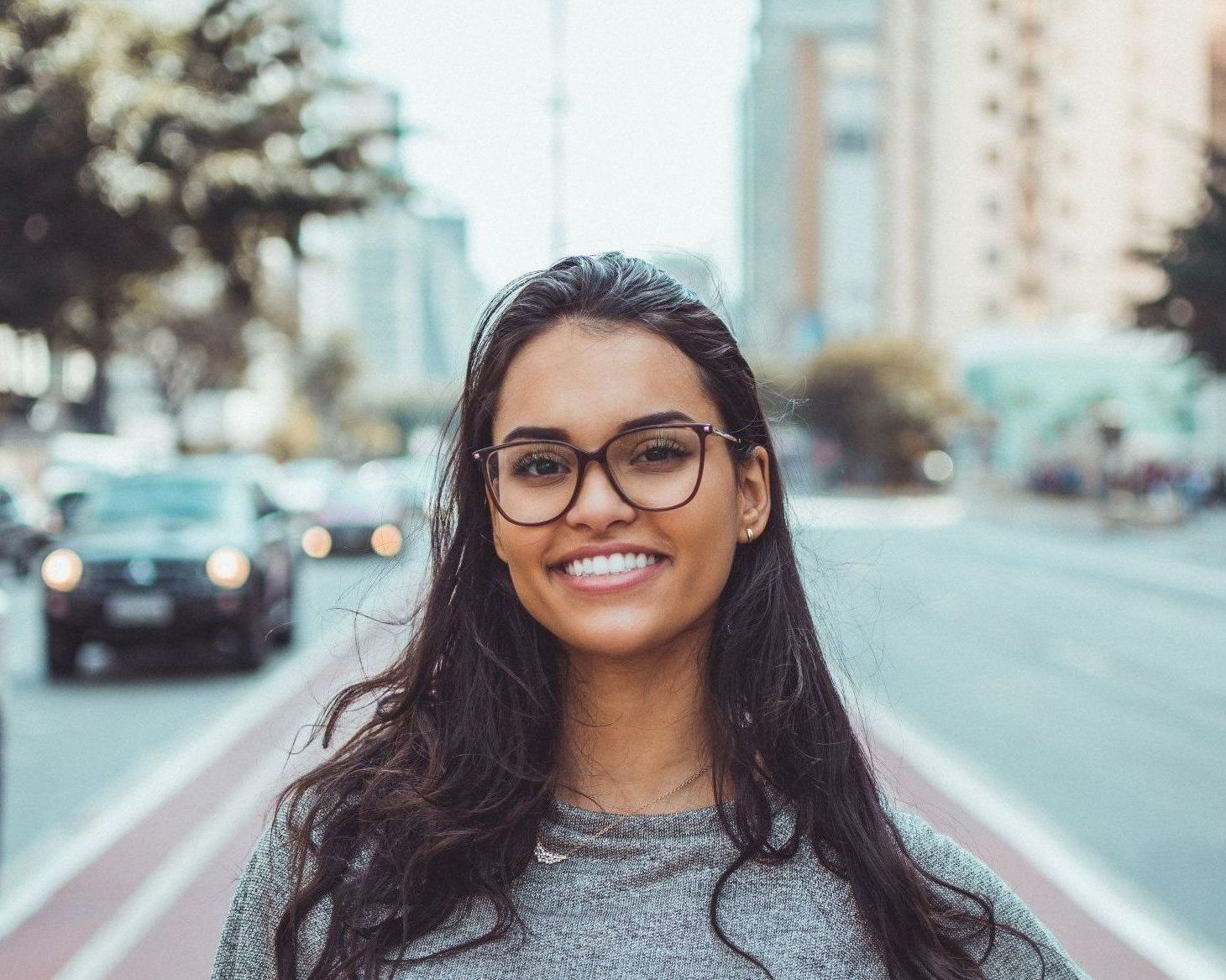 A woman wearing glasses is smiling in front of a city street.