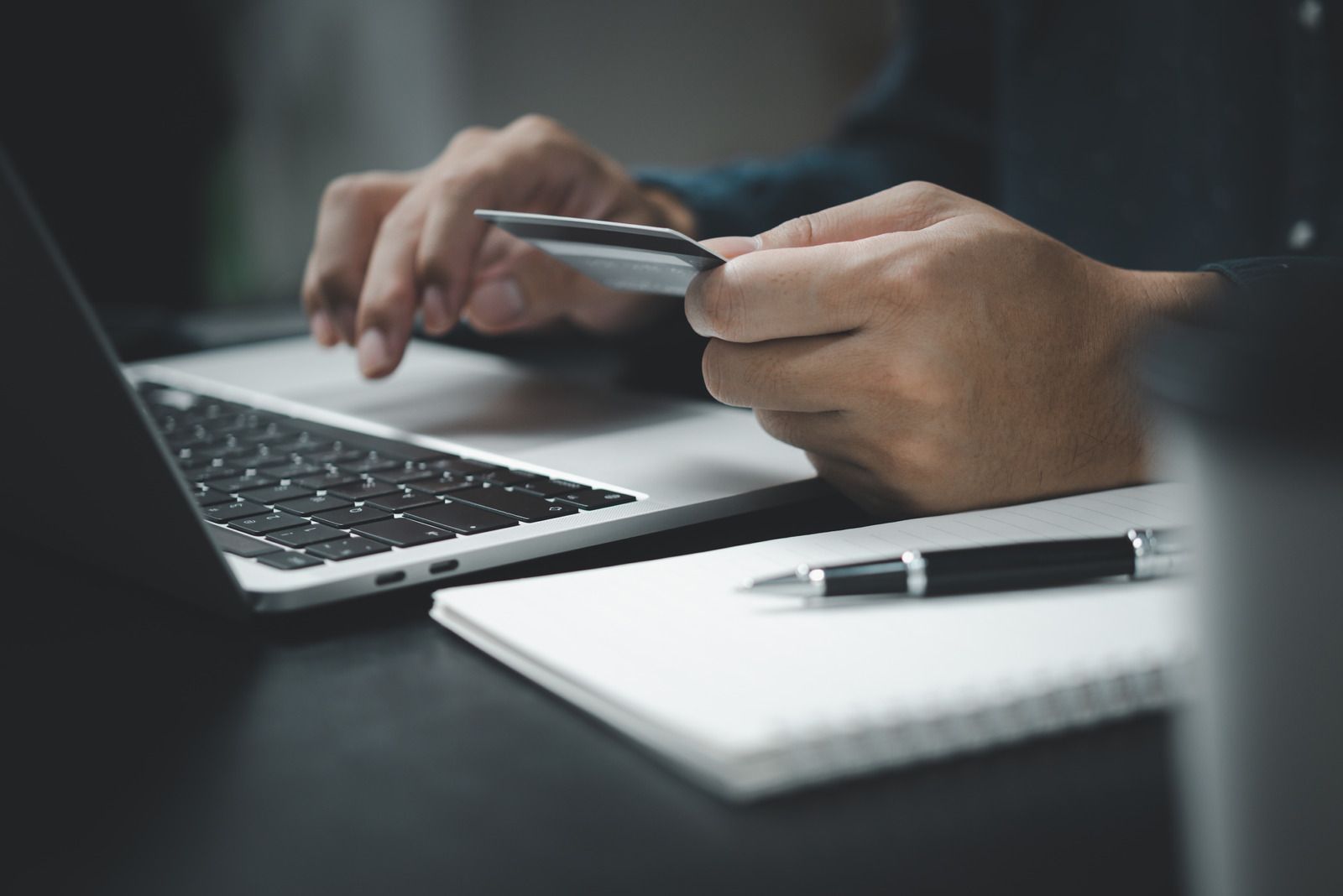 Hands holding a credit card over a laptop, with a notebook and pen on a desk