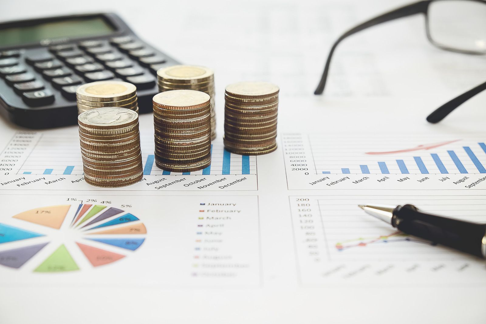 Stacks of coins on business charts with a calculator, pen, and eyeglasses on a desk