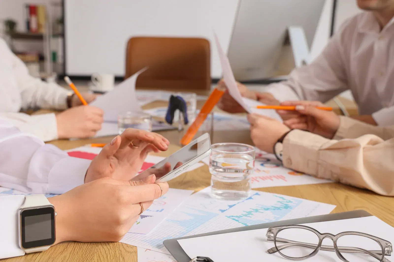 Hands in a business meeting reviewing charts and notes at a table with coffee, glasses, and a smartwatch.