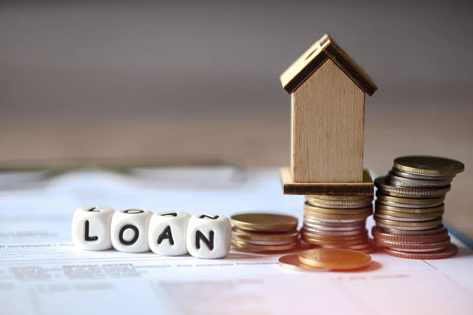 Wooden house model on stacked coins beside letter blocks spelling “LOAN” on a financial document