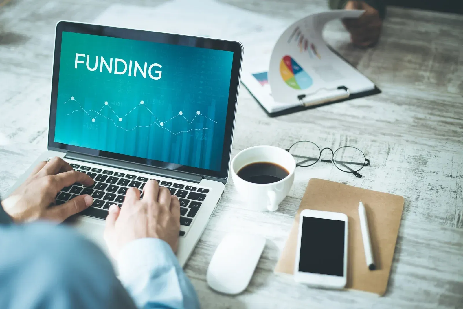 Hands typing on laptop displaying “FUNDING,” with coffee, phone, glasses, and notebook on desk