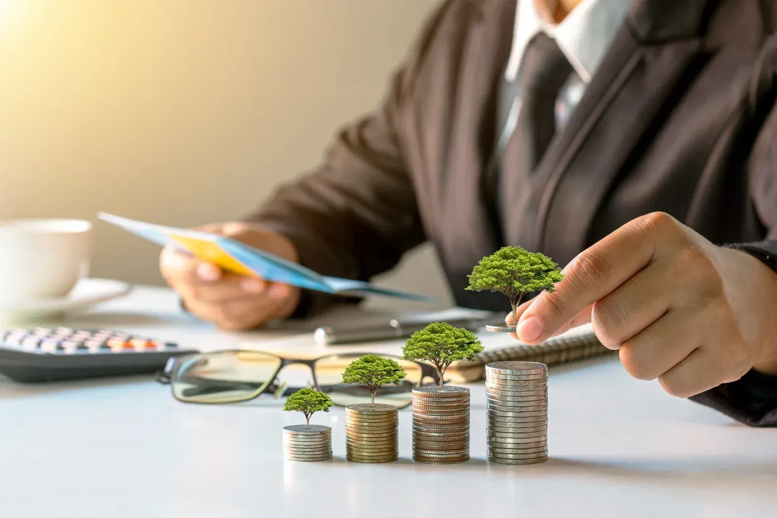 Businessperson stacking coin towers with small trees, symbolizing green investment and growth.