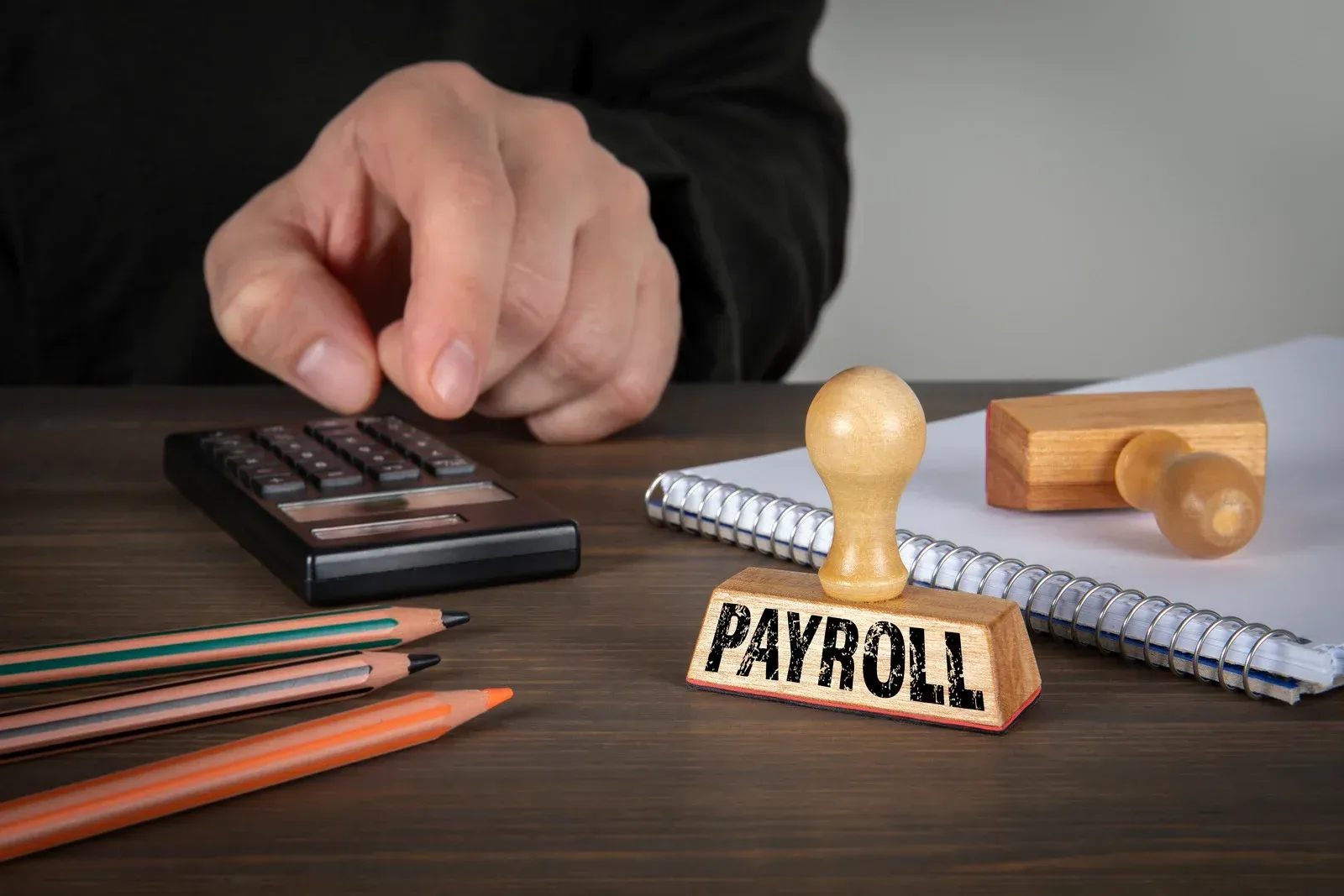 Hand pressing calculator beside rubber stamp labeled “PAYROLL” on a desk with notebooks and pencils