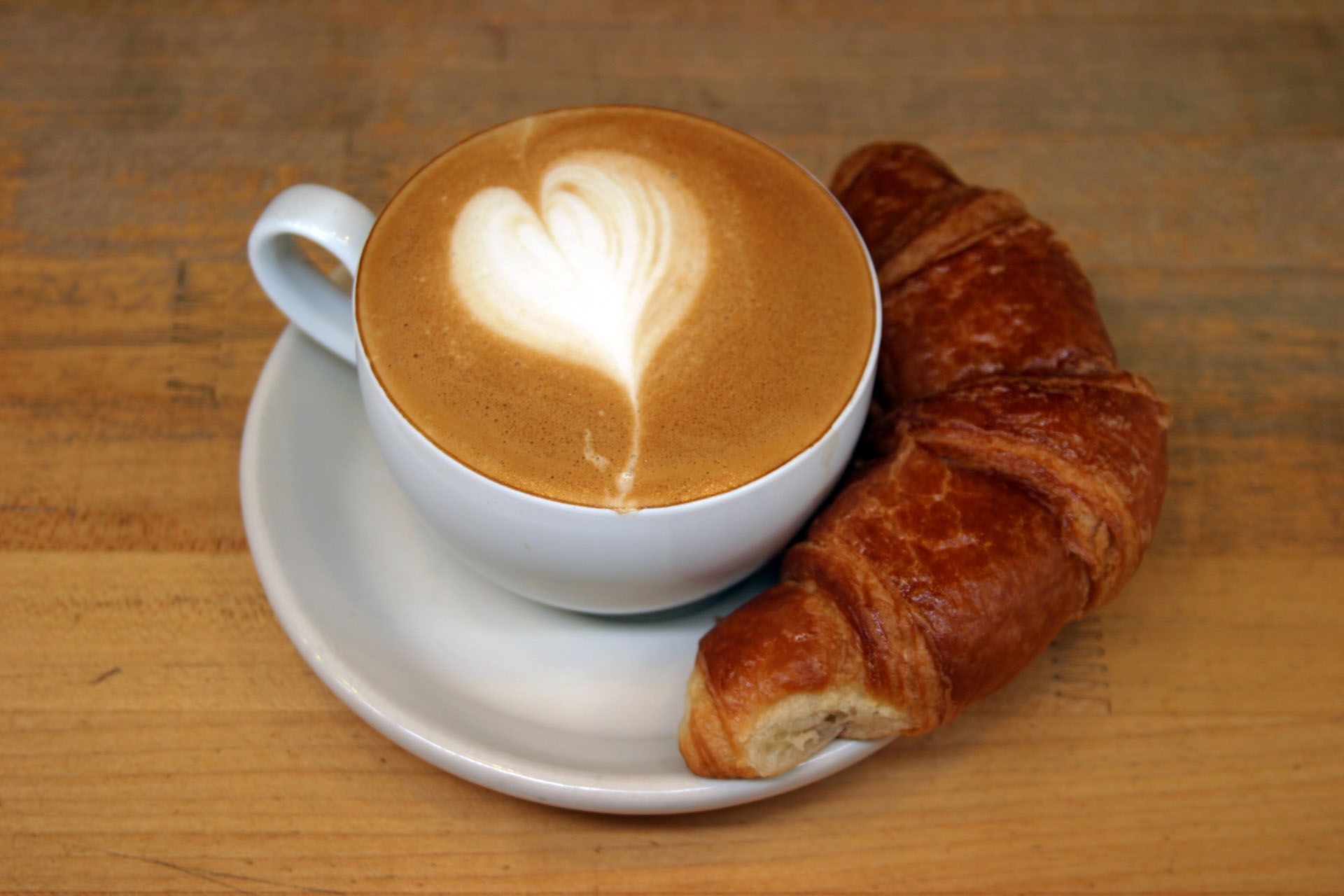 A cup of cappuccino next to a croissant on a wooden table