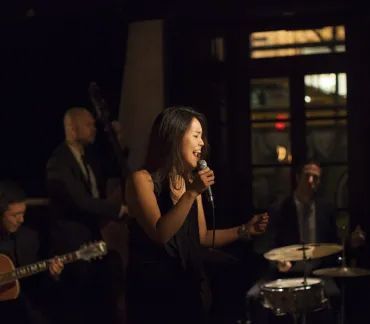 Singer performing on a dimly lit stage with a microphone, guitarist and drummer in the background.