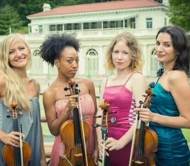 Four women holding violins pose outdoors in front of a building and greenery.