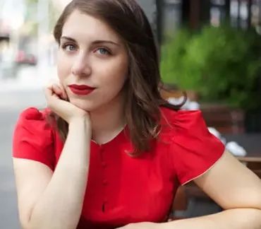 Person in a red blouse resting her chin on her hand at an outdoor café, looking at the camera.