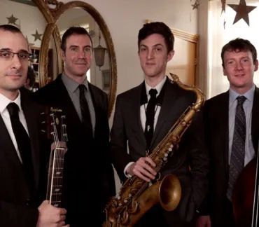 Four men in suits holding musical instruments pose indoors in a formal setting.