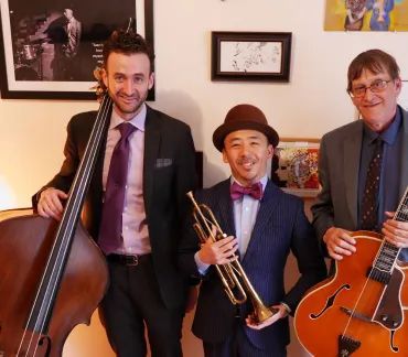 Three musicians posing indoors with a double bass, trumpet, and guitar, smiling in formal attire